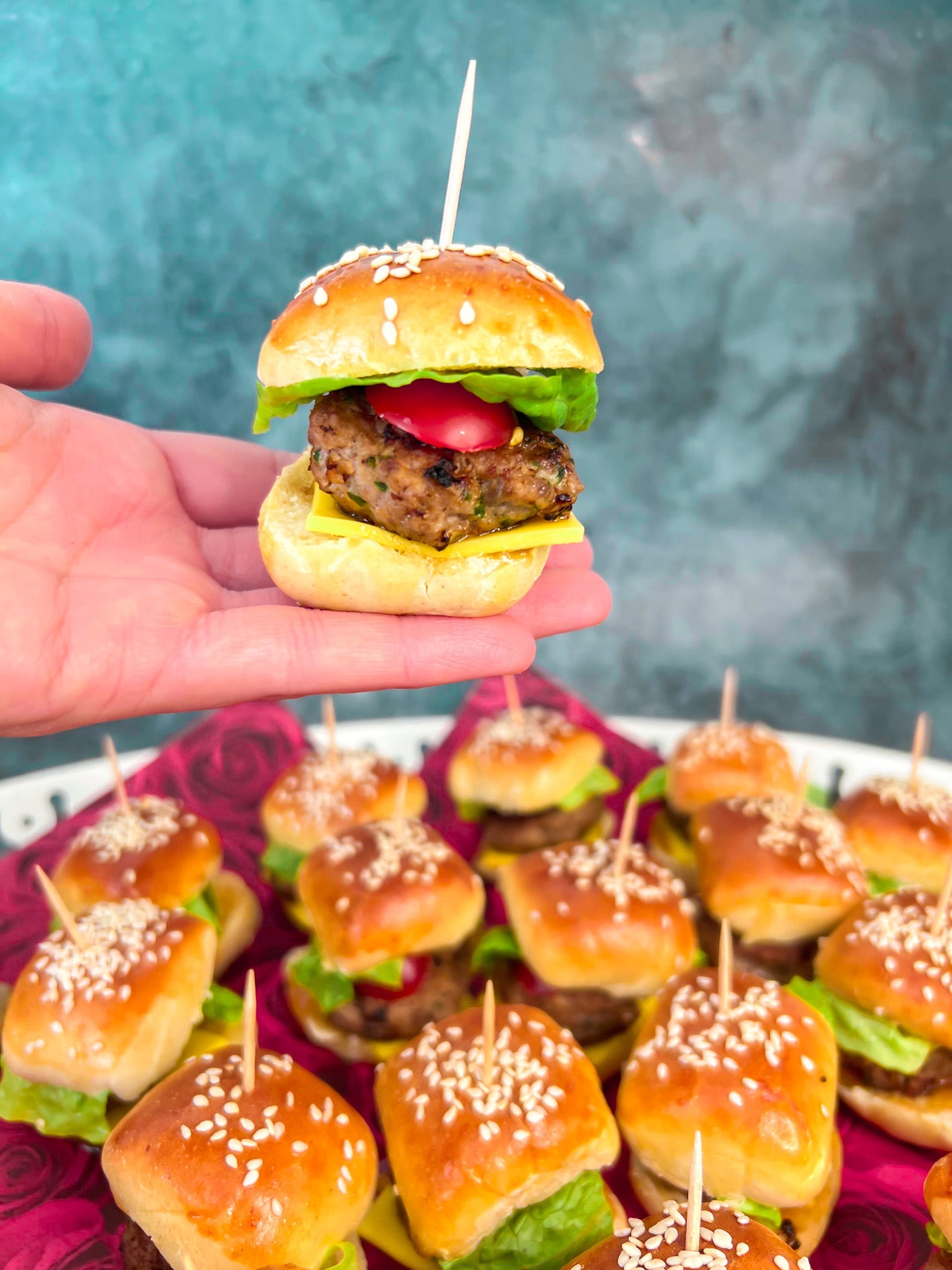 Mini-Burger mit Sesambrötchen, saftigem Hackfleisch-Patty, Salat, Cherrytomate und Käse, fixiert mit einem Zahnstocher und serviert auf einem Teller als Fingerfood.