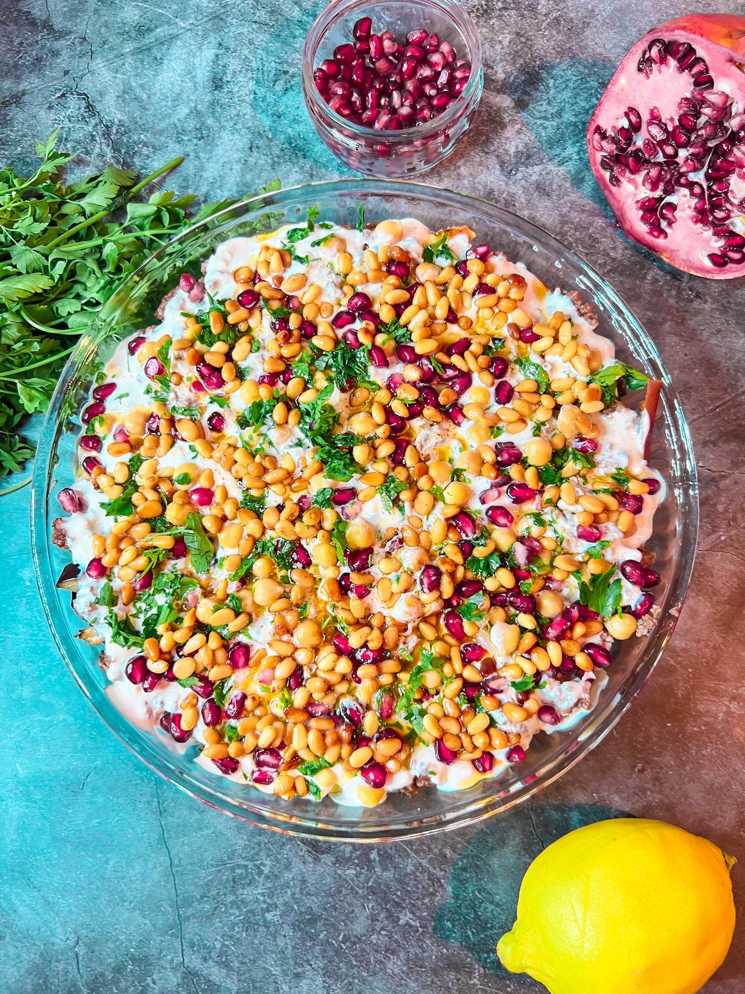 Fatteh mit Auberginen, Kichererbsen, Hackfleisch und Joghurtsoße, geschichtet mit frittiertem Fladenbrot, garniert mit Pinienkernen, Granatapfelkernen und Petersilie.