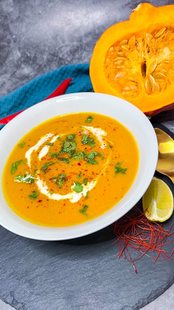 Eine Schale asiatische Kürbissuppe, garniert mit gehacktem Koriander und einem Klecks Kokosmilch, serviert auf einem rustikalen Tisch.